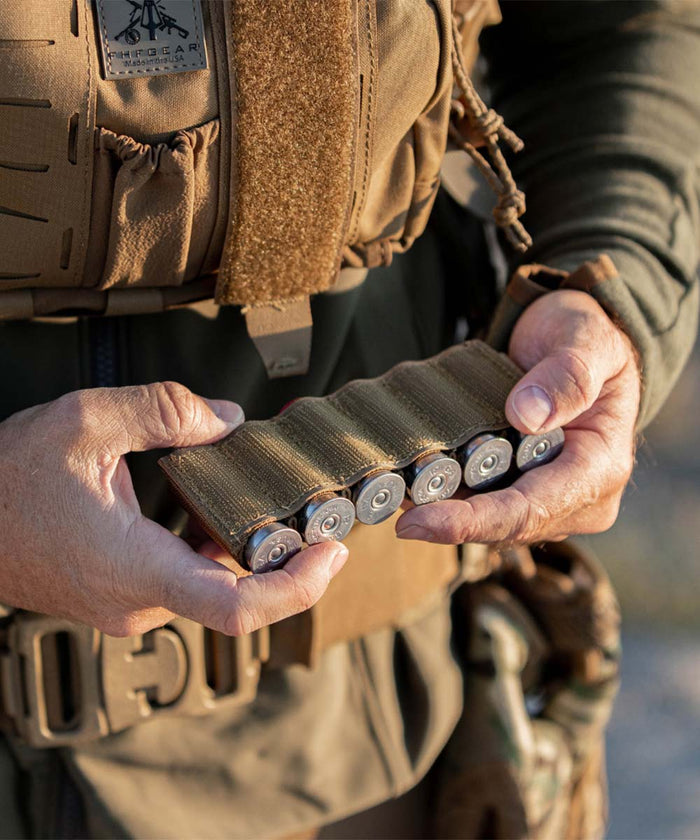 Tan shotgun shell card holding five silver shells, gripped by hands against a tactical vest.