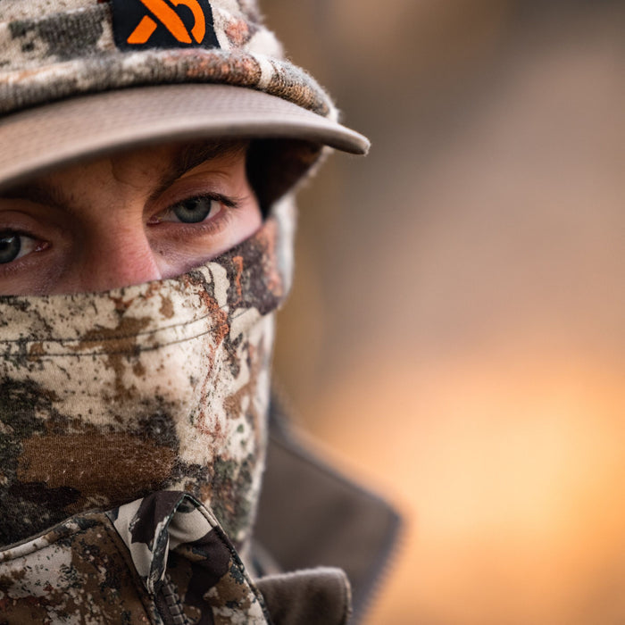 Camouflage hunting balaclava, hat with an orange logo, and jacket worn by a person, with only their eyes visible.