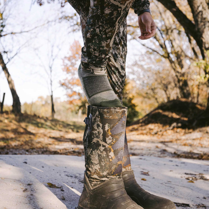 A grey and olive green patterned sock is pulled partially out of a muddy camouflage rubber boot worn with camouflage pants.