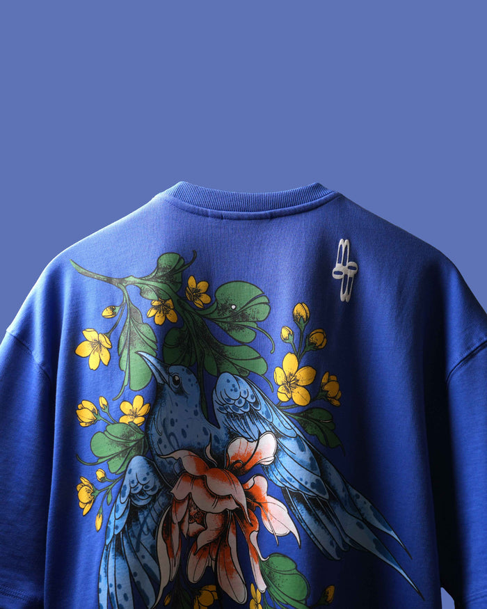 A royal blue crewneck t-shirt featuring a large graphic print on the back with blue birds, yellow flowers, and green leaves, shown against a solid blue background.