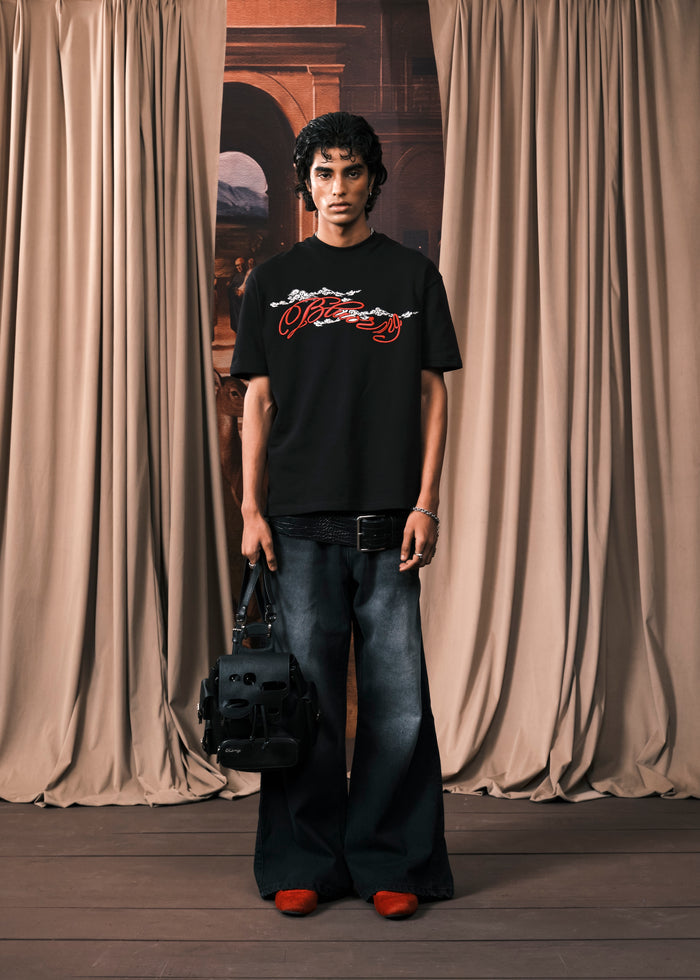 A model stands in front of beige curtains wearing a black graphic t-shirt with a red and white 'Pleeszy' logo, wide-leg dark jeans, and red shoes, holding a black shoulder bag.