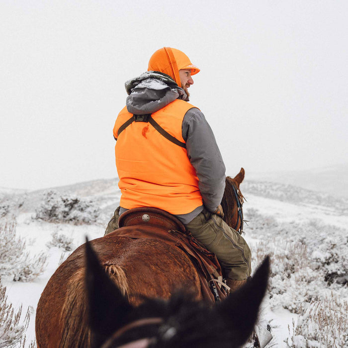 Blaze orange hunting vest with black straps and matching hooded cap worn by a person on horseback in a snowy landscape.