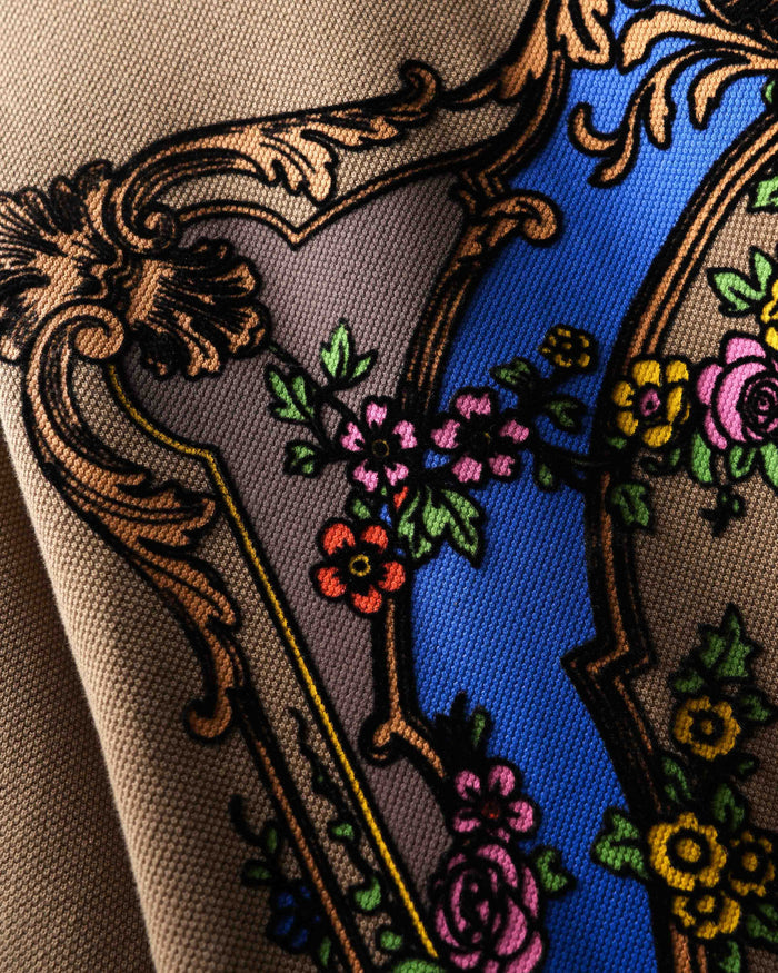 Close-up of the back of a beige polo t-shirt featuring a large ornate baroque-style graphic with a blue arch, gold scrollwork, and floral details.