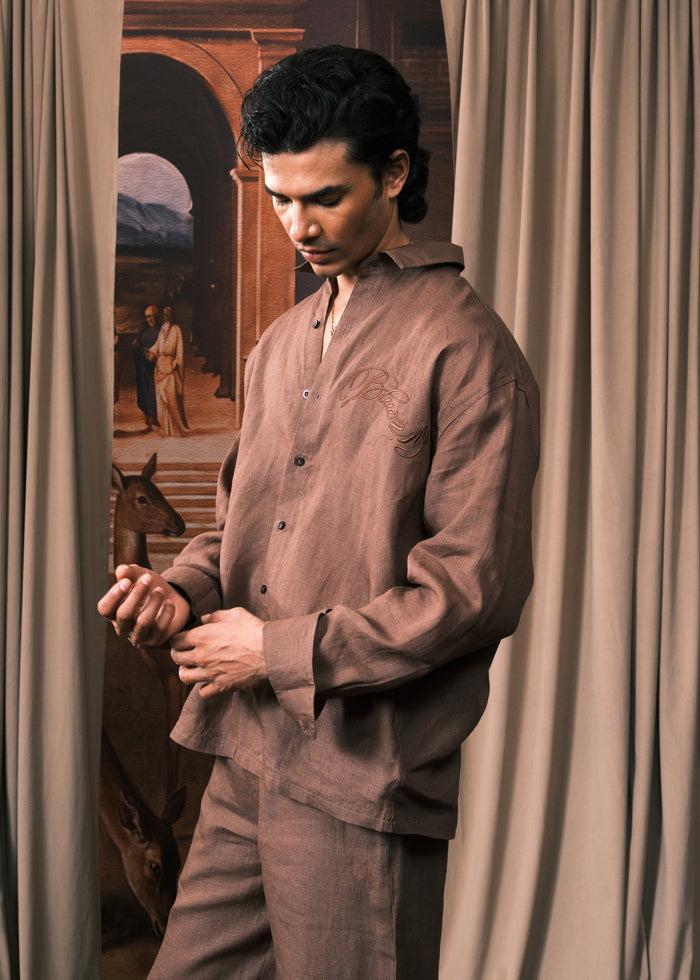 A man wearing a matching brown linen pajama set consisting of a long-sleeve button-up shirt and trousers, standing in front of beige curtains with a vintage painting in the background.