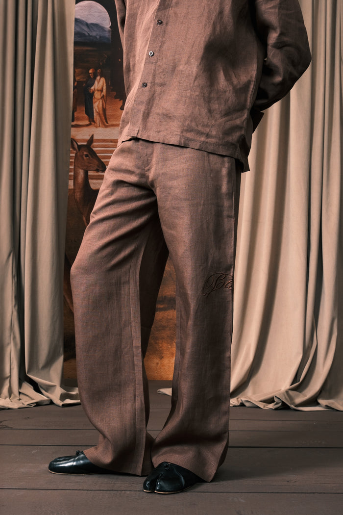 A person wearing a matching brown linen pajama set with a button-up shirt and wide-leg pants, paired with black loafers, standing in front of draped curtains.