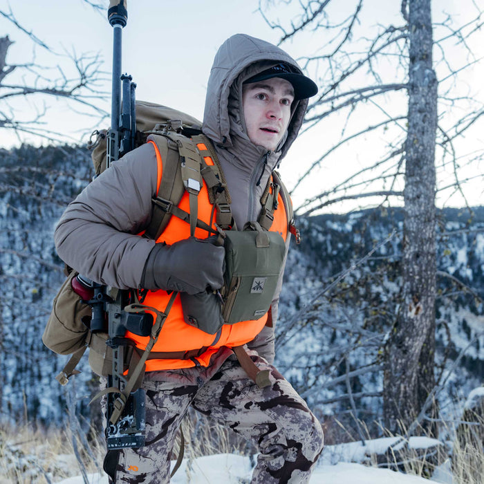 Man in tan hooded jacket, orange safety vest, and camo pants, carrying a rifle strapped to a backpack and wearing an olive chest rig.
