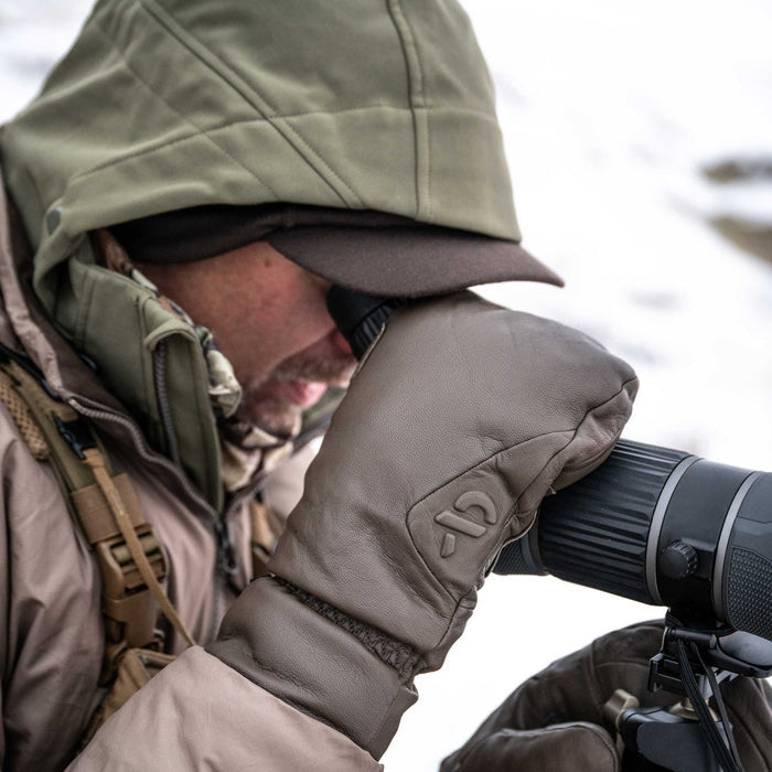 A person wearing dark brown leather mittens with an embossed logo is looking through a spotting scope in a snowy outdoor setting.