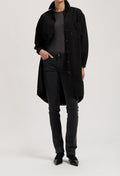 Model wearing a long black button-up coat over a gray top, paired with fitted black jeans and pointed black shoes.