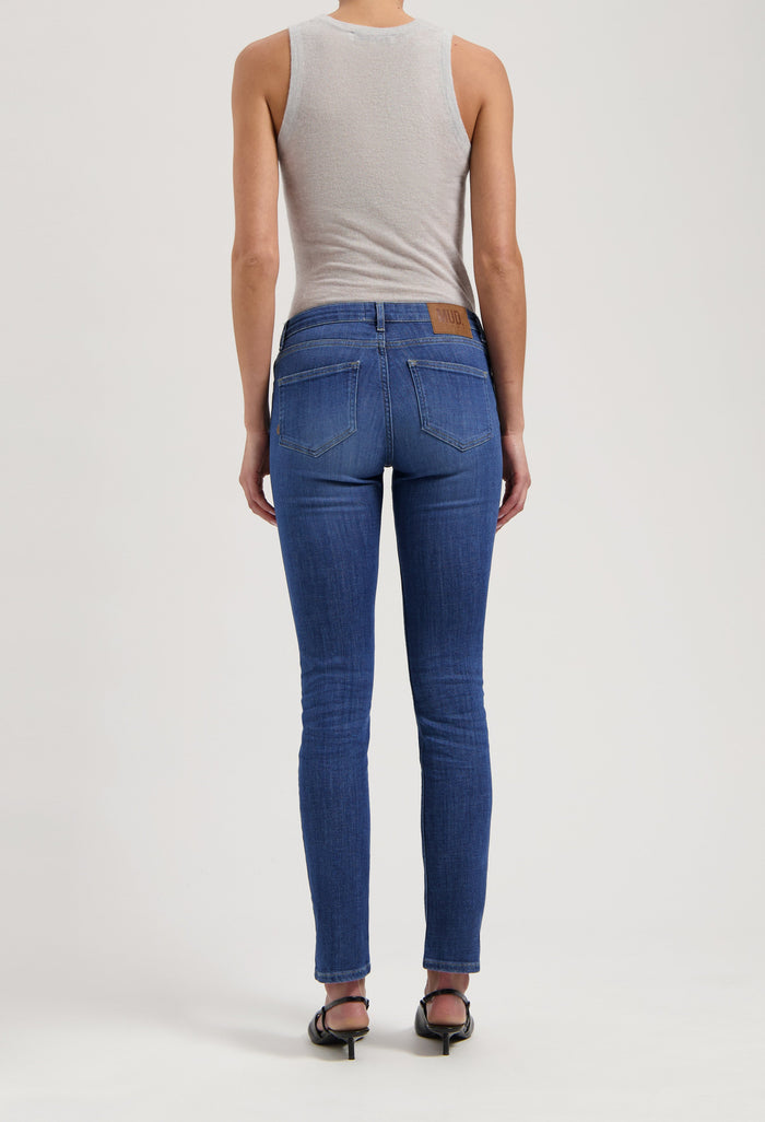 Back view of slim-fit, sustainable blue jeans by MUD Jeans, paired with a light gray sleeveless top and black sandals.