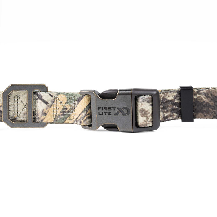 A close-up of a camouflage dog collar with a dark gray metal quick-release buckle. The collar features a woodland camo pattern in shades of green, brown, and beige, with the brand name 'FIRST LITE' clearly visible on the buckle.