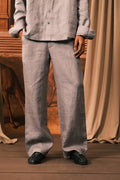 A person wearing a light gray linen button-up shirt and matching wide-leg trousers, standing on a wooden floor with draped beige fabric and a rustic wooden backdrop.