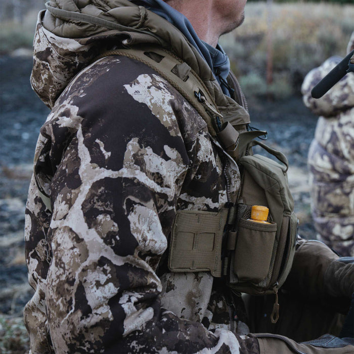 A person wears a brown, tan, and black camouflage hunting jacket with a padded hood and an olive green chest rig featuring a yellow-capped bottle.