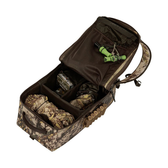Camouflage hunting blind bag open, showing organized internal compartments with two green duck calls, a box of shotgun shells, and camouflage gloves, plus full external shell loops.