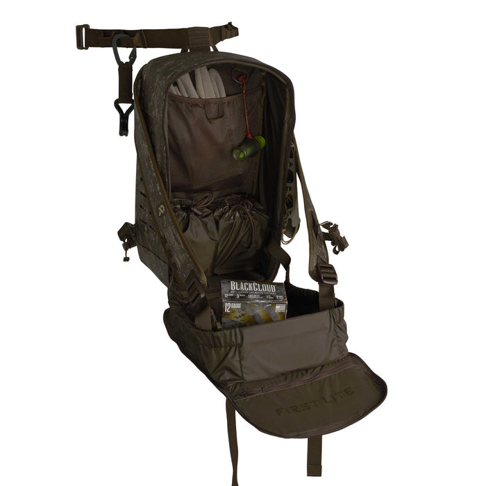 A dark camouflage First Lite hunting backpack is open, revealing internal storage including two drawstring pouches, a duck call, and a box of 12 gauge ammunition.