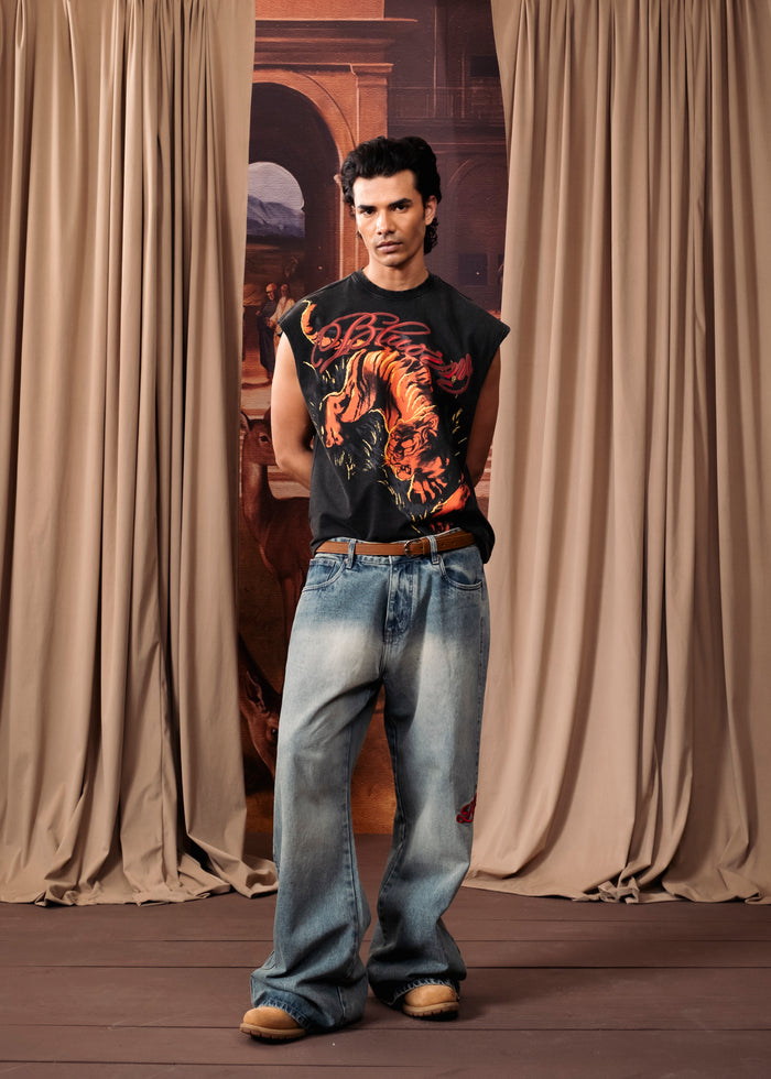 A man modeling a black sleeveless graphic t-shirt with a red and orange tiger design, paired with light blue jeans and brown shoes, standing in front of draped beige curtains.