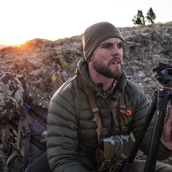 A man outdoors wears an olive green beanie and a quilted olive green puffer jacket with an orange chest logo.