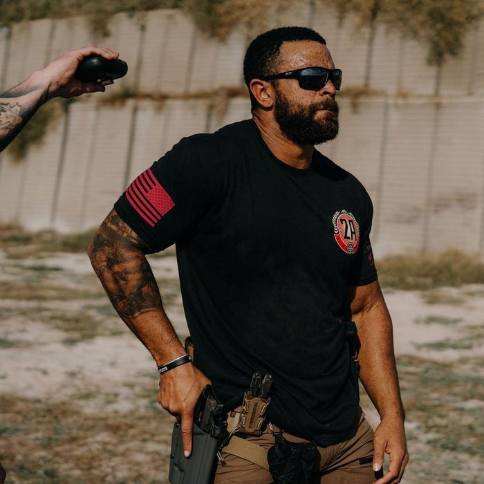 Man wearing a black t-shirt with red and white designs, holding a rifle outdoors.