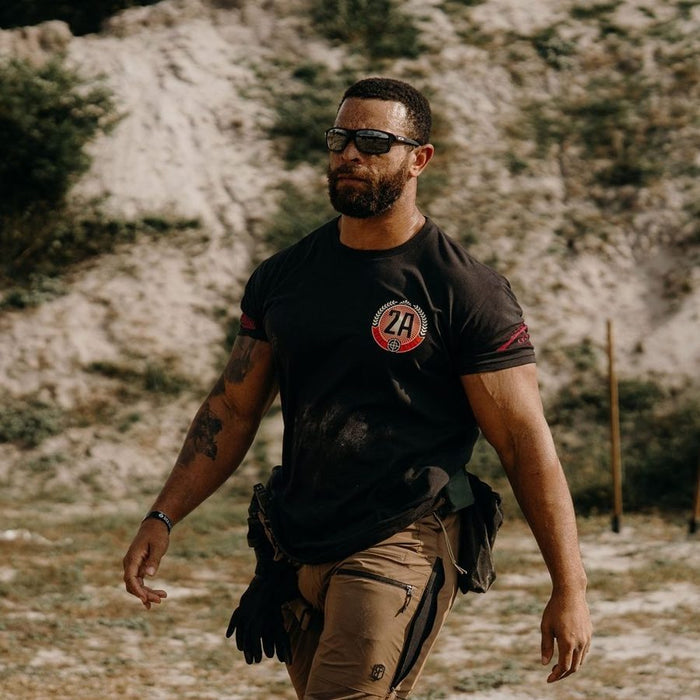 Man wearing sunglasses and a black t-shirt with a logo, standing in a natural setting.