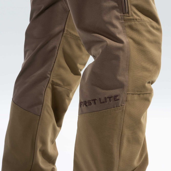 Close-up of a person's lower legs wearing First Lite outdoor pants in a brown and khaki color-blocked design, with the brand name 'FIRST LITE' embroidered on the side.