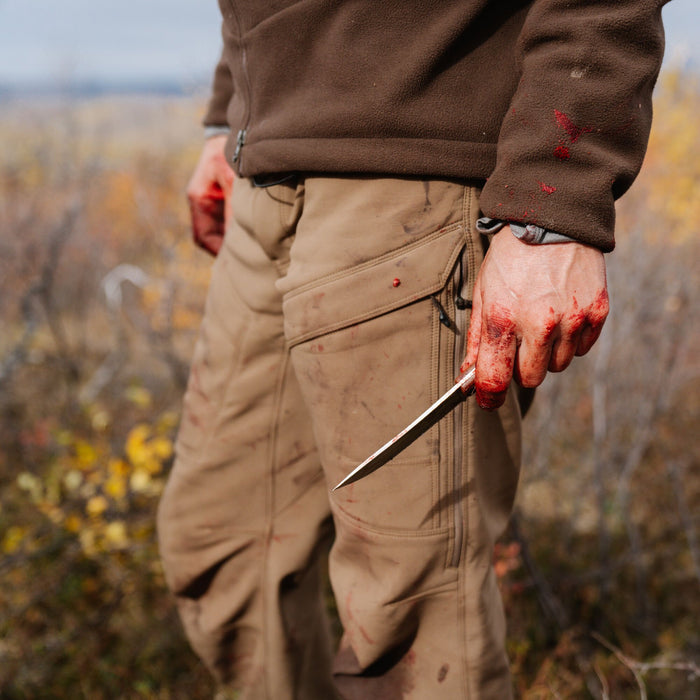 A person wears a blood-stained brown fleece jacket and tan cargo pants, holding a bloody silver knife.