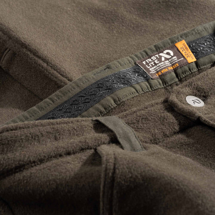 Close-up of a green wool hunting pant showing the inner waistband, a black silicon strip,, and a sewn-in orange and white 'FIRST LITE' brand tag.