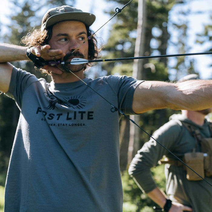 Slate gray short-sleeve t-shirt featuring a "FIRST LITE" sun logo and "GO FURTHER. STAY LONGER." text on the chest.
