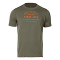 An olive green t-shirt features orange text on the front saying 'Born in the Wild of Idaho, USA First Lite Go Farther | Stay Longer Since Before Smart Phones' and a small brown tag on the lower right hem.