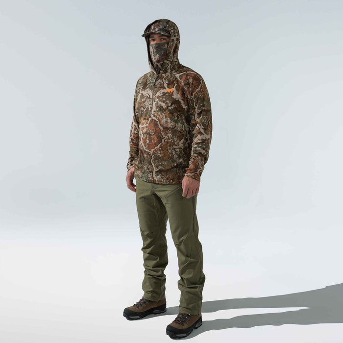 Man wears a brown and green camouflage hooded 1/2-zip top with integrated face mask, visor, and orange logo, olive pants, and brown hiking boots.