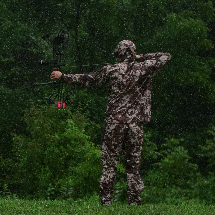 A person in a brown, gray, and white camouflage hunting suit with a hooded jacket aims a compound bow in the rain.