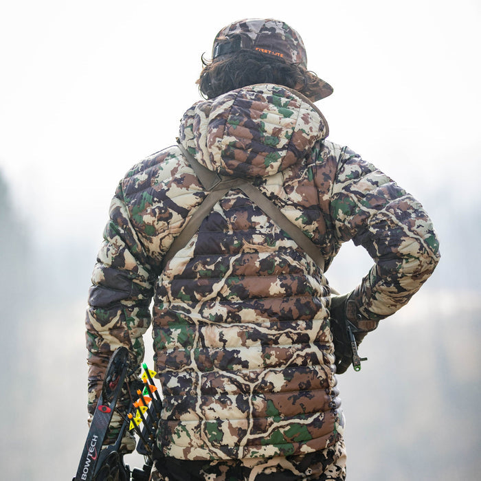 A person viewed from behind wears a First Lite Fusion camouflage quilted puffer jacket and hat, holding a Bowtech compound bow with arrows.