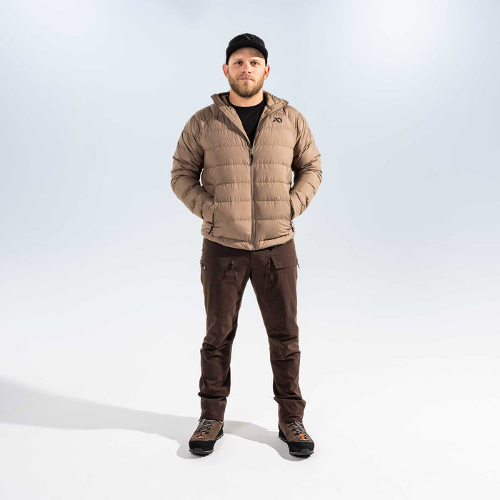 A man wearing a light brown puffer jacket, dark brown pants, and a black cap, standing against a light gray studio background.