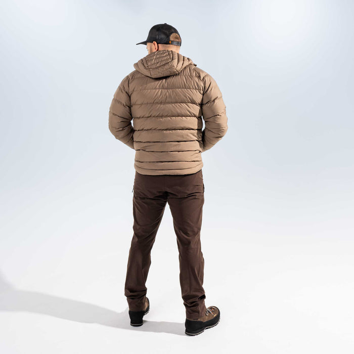 A man viewed from behind wearing a tan hooded puffer jacket, dark brown pants, and a black baseball cap against a light gray studio background.