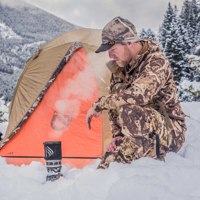 A man in camouflage crouches in deep snow, cooking on a black Jetboil stove with steam rising, next to an orange and tan Nemo tent in a winter mountain setting.