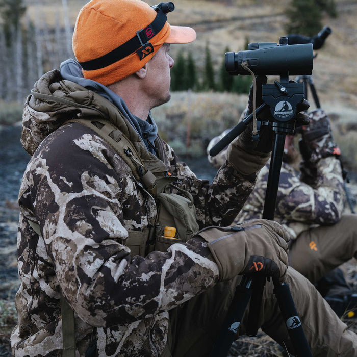 A person in an orange beanie with a headlamp and camouflage jacket holds a black spotting scope on a black Outdoorsmans tripod.