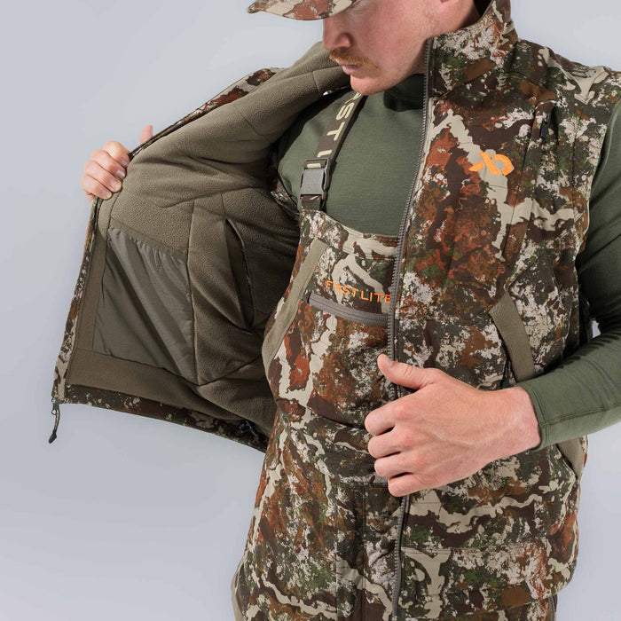 Man in a multi-color camouflage vest with an orange logo and bibs, revealing the vest's olive green fleece interior with an internal mesh pocket, and "FIRST LITE" text on the bib pocket.