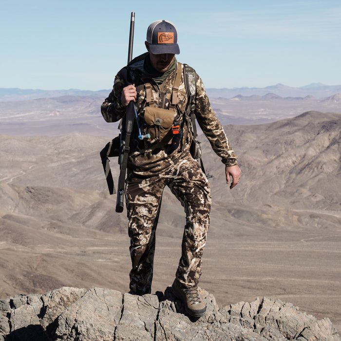Man in brown and tan desert camouflage hunting outfit, including long-sleeved shirt and pants, stands on a rocky outcrop with a rifle.