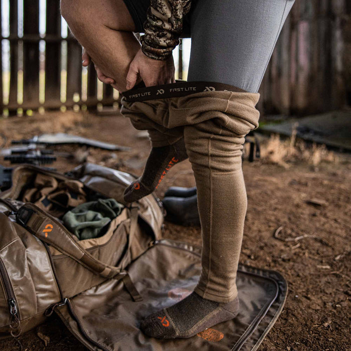 A person pulls on brown First Lite base layer pants with a visible branded waistband, wearing dark socks with orange logos.