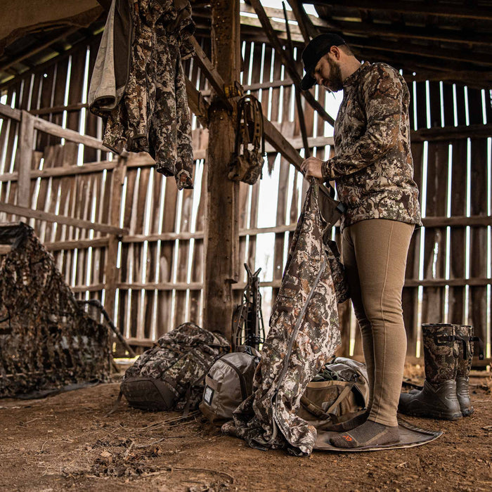Man inspects brown and green camouflage hunting outerwear with full-length zippers inside a rustic barn filled with hunting equipment.
