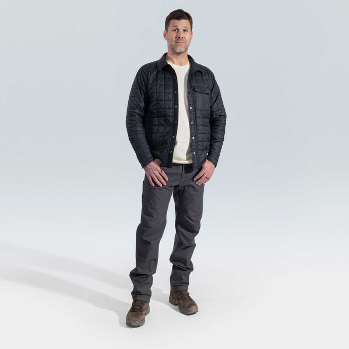 Man wearing a dark quilted jacket, white shirt, gray pants, and brown boots on a light gray background