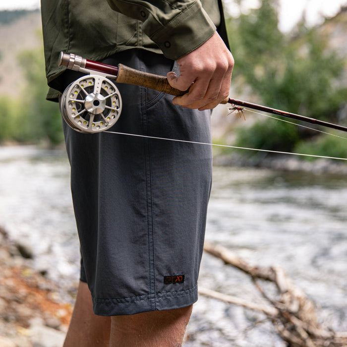 A person wears dark gray shorts with a small orange logo on the right leg, holding a fishing rod by a river.