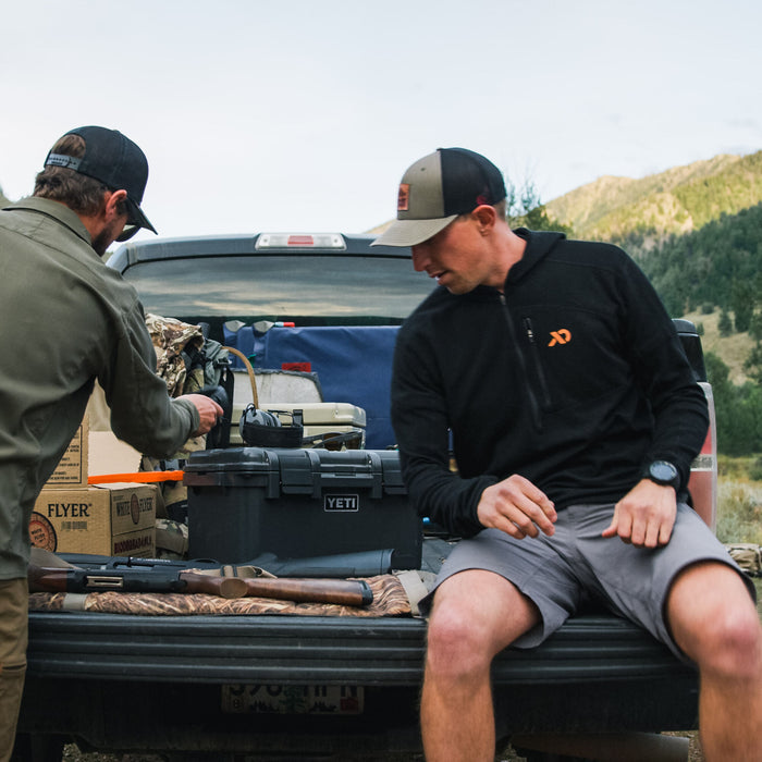 Two men prepare gear in a truck bed, featuring a black Yeti gear case, a shotgun with a wooden stock, and brown White Flyer clay target boxes.