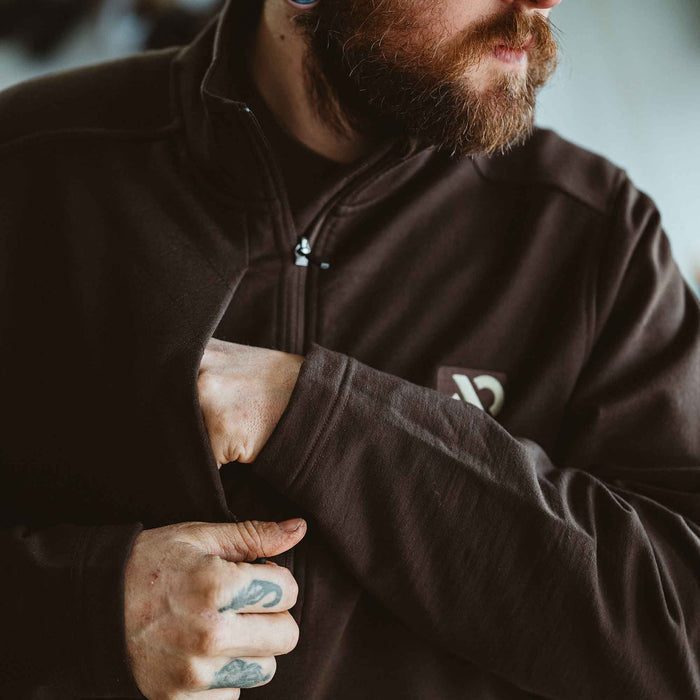 Dark brown full-zip jacket with a subtle chest logo, worn by a person reaching into a side pocket.