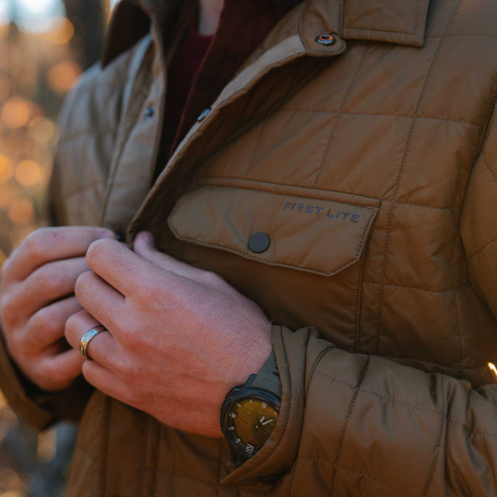 Close-up of a person buttoning a brown quilted jacket featuring a chest pocket with the "FIRST LITE" logo and snap buttons.