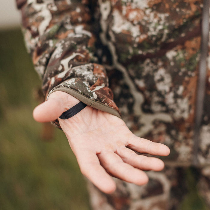 Close-up of a person's hand extended, palm up, wearing a brown and green camouflage jacket with a black thumb loop at the cuff.