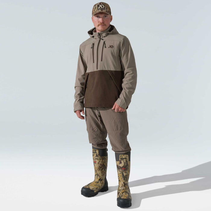 Man in a two-tone tan and dark brown hooded pullover, tan pants, tall camouflage rubber boots, and a camouflage cap.