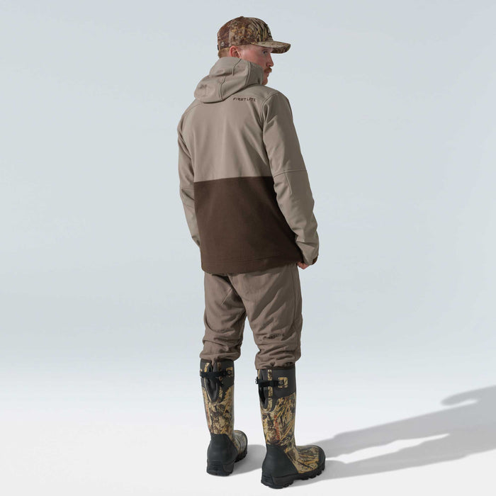 A man wears a two-tone tan and dark brown hooded jacket with 'FIRST LITE' logo, brown pants, a camouflage cap, and tall camouflage hunting boots, viewed from behind.