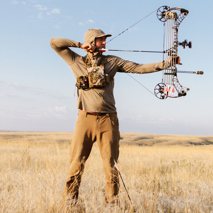 Hunter in tan hooded long-sleeve shirt and pants, with a camo chest rig, aiming a compound bow with a pink-fletched arrow in a field.