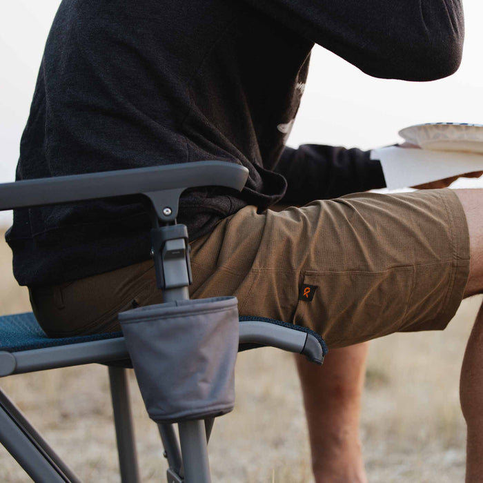 Earthy brown textured shorts with a small orange loop logo tag on the right thigh pocket, worn by a person sitting outdoors.