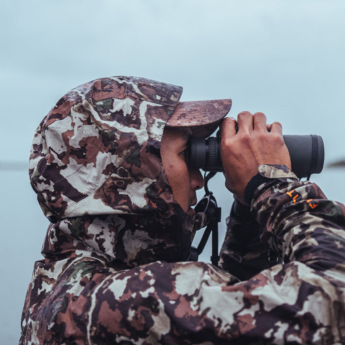 Hooded earth-toned camouflage rain jacket with visible water droplets and an orange First Lite logo on the sleeve.