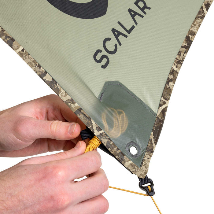 A person's hands adjust a sage-green Scalar tarp with camouflage edges, a yellow rope threaded through a black cord lock, and a transparent pocket revealing a coiled rope.